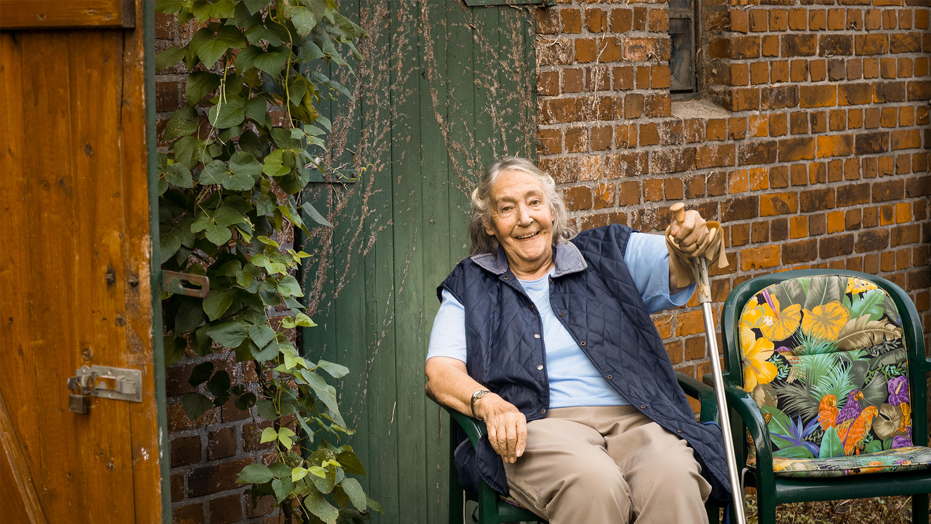 Ältere Frau sitzt in ihrem Garten und lächelt in die Kamera